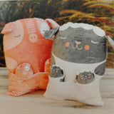 Two plush toys, one orange hippo and one gray cat, on a wooden surface with a blurred natural background.