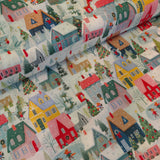 A close-up of a festive Christmas-themed fabric or wallpaper print featuring a snowy village with colorful houses, trees, and small figures on a white background.